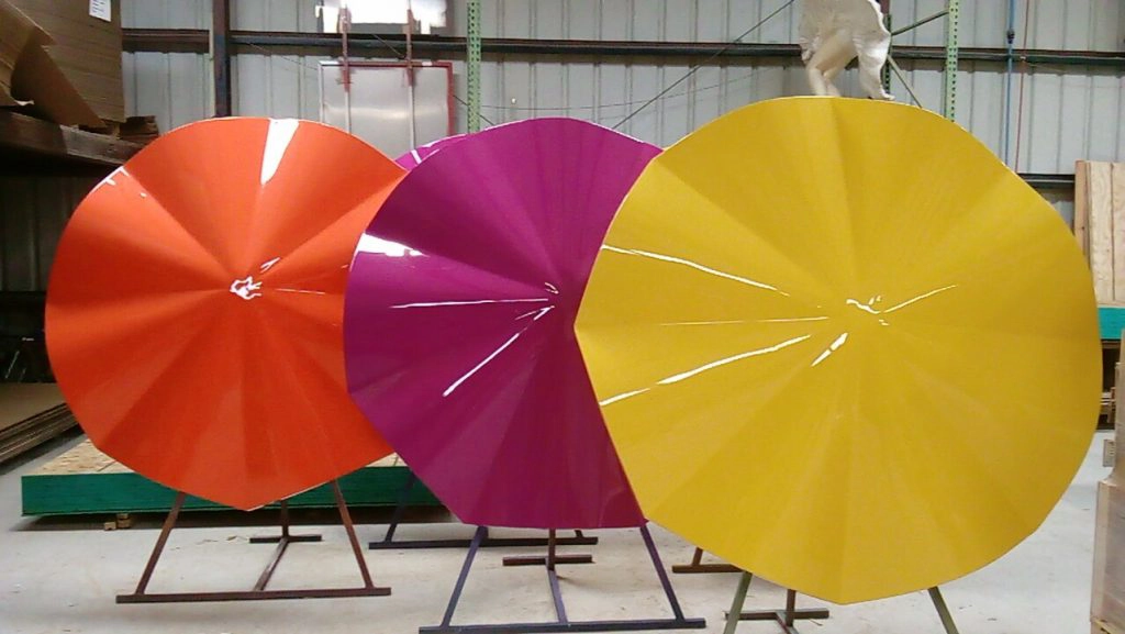 Three glossy fiberglass hard-top umbrella canopies in orange, purple, and yellow displayed on support stands inside a warehouse